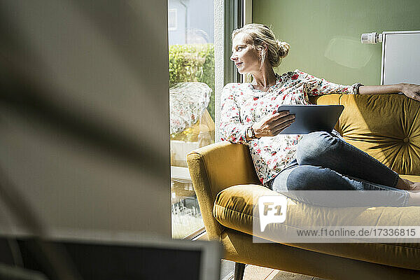 Woman with digital tablet looking through window while sitting on sofa in living room
