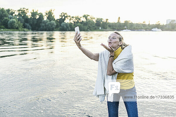 Frau bläst Kuss während der Aufnahme von Selfie durch Smartphone am Flussufer