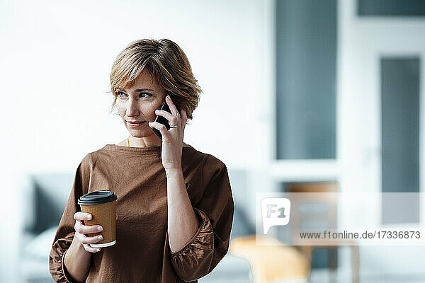 Geschäftsfrau mit Kaffeetasse  die im Büro mit einem Mobiltelefon spricht