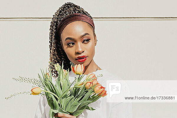 Beautiful young woman holding flower bouquet while looking away in front of white wall on sunny day