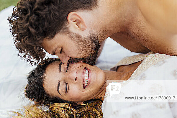 Zärtlicher Mann küsst glückliche Freundin mit geschlossenen Augen auf Decke liegend