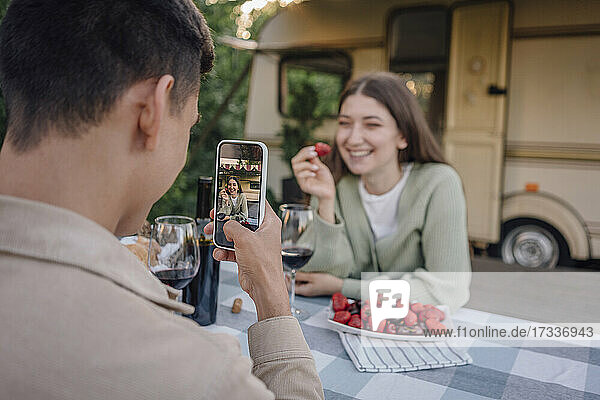 Young man photographing girlfriend holding strawberry through smart phone sitting at table