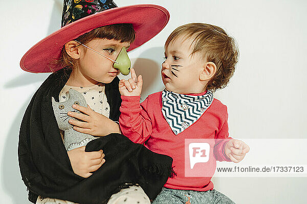 Girl in Halloween costume looking at sister touching artificial nose by wall