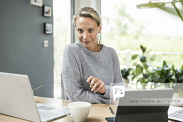 Businesswoman holding eyeglasses while sitting by laptop and digital tablet at home
