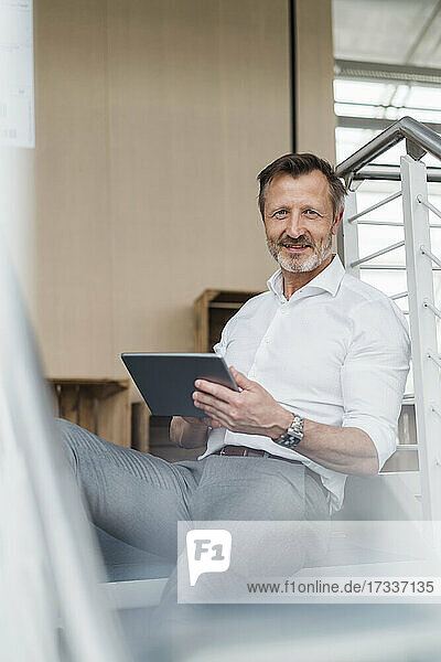Male professional holding digital tablet while sitting on steps in office