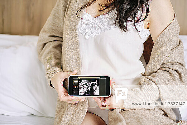 Schwangere Frau zeigt Ultraschallbild auf Smartphone zu Hause