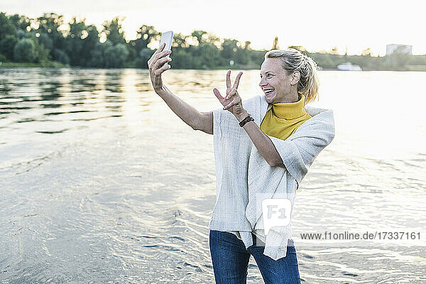 Lächelnde Frau gestikuliert Friedenszeichen bei der Aufnahme von Selfie durch Smartphone am Flussufer