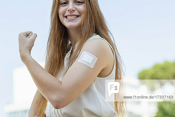 Lächelnde rothaarige Geschäftsfrau  die nach einer Impfung während der Pandemie ihre Muskeln spielen lässt