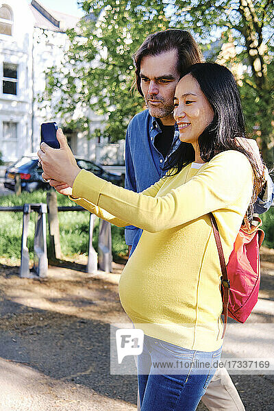 Smiling pregnant woman taking selfie through mobile phone with husband at public park