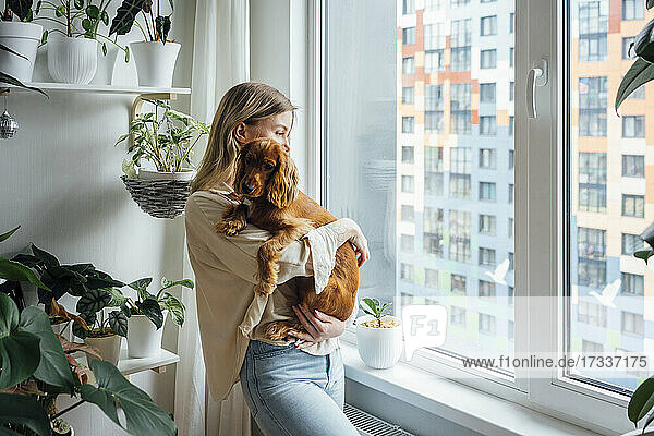 Blonde Frau mit Hund schaut weg  während sie zu Hause am Fenster steht
