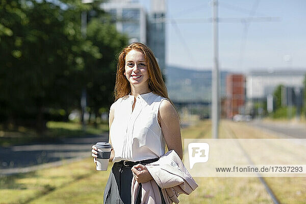 Lächelnde Freiberuflerin mit Einweg-Kaffeebecher an einem Eisenbahnregal stehend