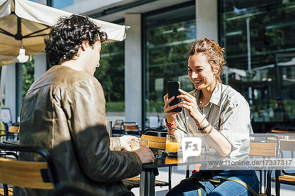 Lächelnde Frau  die ihren Freund mit dem Smartphone in einem Straßencafé fotografiert