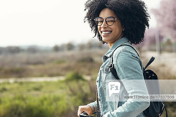 Fröhliche Frau mit Brille lächelt  während sie mit einem elektrischen Tretroller unterwegs ist