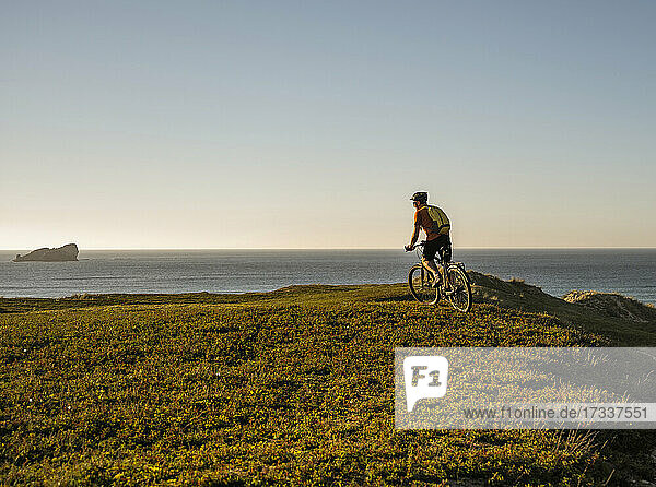 Männlicher Sportler mit Rucksack beim Radfahren auf grünem Gras bei Sonnenuntergang