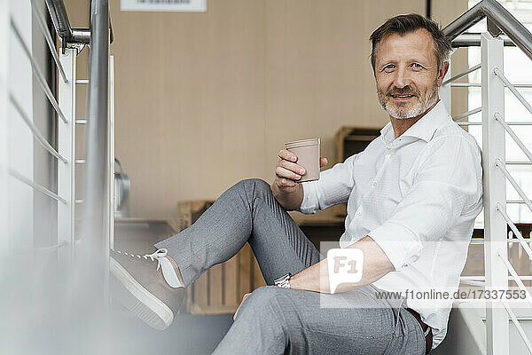 Lächelnder Geschäftsmann hält Kaffeetasse  während er auf einer Treppe sitzt