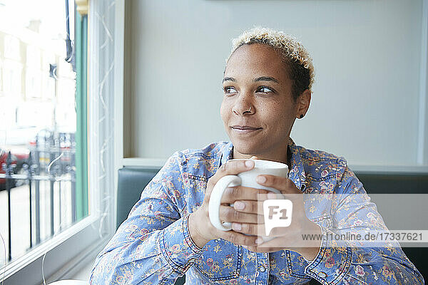 Mittlere erwachsene Frau  die durch ein Fenster schaut und eine Kaffeetasse hält  während sie in einem Café sitzt