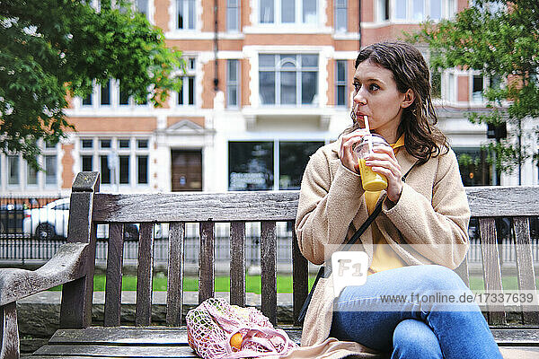 Young woman looking away while drinking smoothie on bench