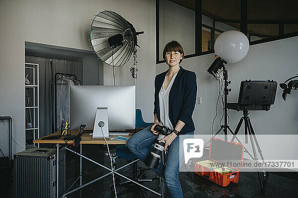 Selbstbewusste Fotografin  die eine Digitalkamera in der Hand hält  während sie in einem Studio auf dem Schreibtisch sitzt