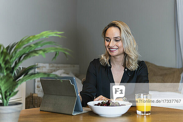 Lächelnde Geschäftsfrau arbeitet an einem Tablet-PC in der Nähe einer Schüssel mit Kirschen und Saft am Schreibtisch im Heimbüro