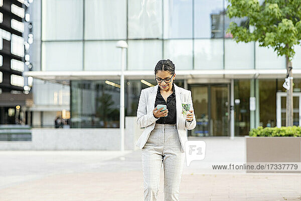 Geschäftsfrau  die ein Smartphone benutzt  während sie vor einem Bürogebäude spazieren geht
