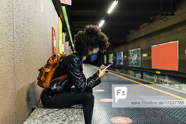 Frau mit Gesichtsschutz  die in einer U-Bahn-Station sitzt und ein Mobiltelefon benutzt