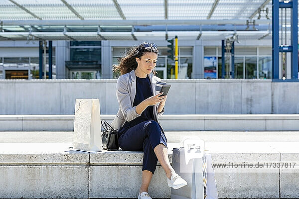 Woman with shopping bags using smart phone while sitting on retaining wall
