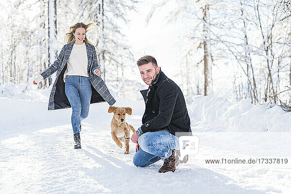 Mann hockt im Schnee  während seine Freundin im Urlaub mit dem Hund im Schnee läuft