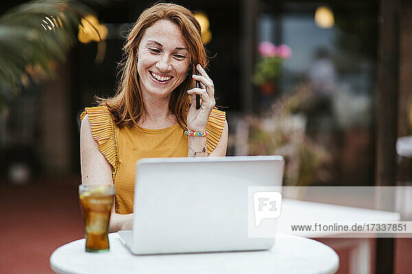 Redhead woman talking on smart phone while using laptop at table
