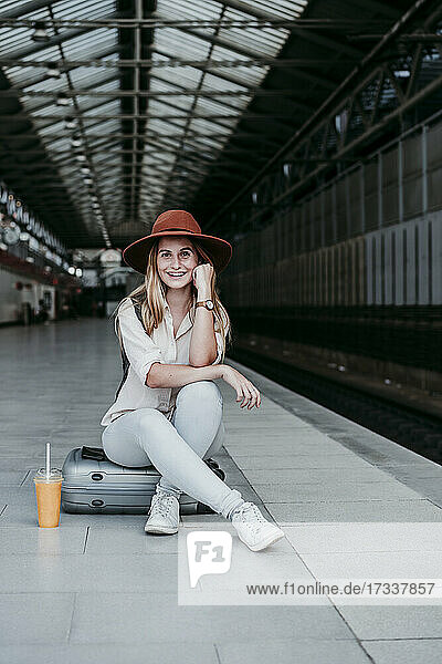 Glückliche Frau mit Hut  die auf einem Gepäckstück auf dem Bahnsteig sitzt