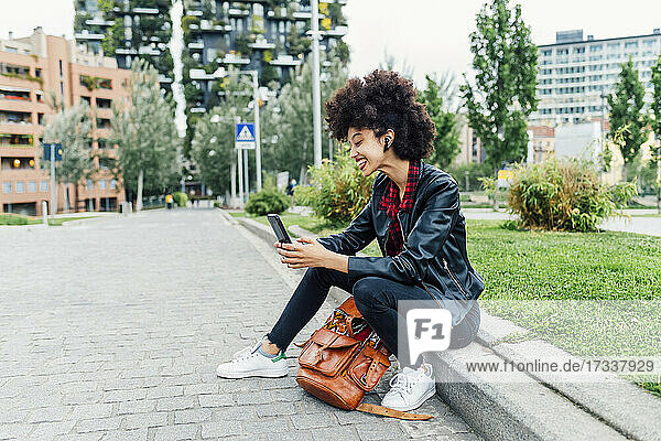 Cheerful woman with wireless in-ear headphones using mobile phone at park