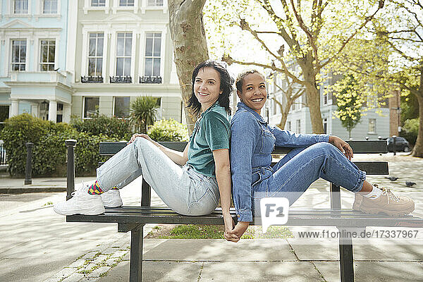 Lächelndes lesbisches Paar hält sich an den Händen und sitzt Rücken an Rücken auf einer Bank