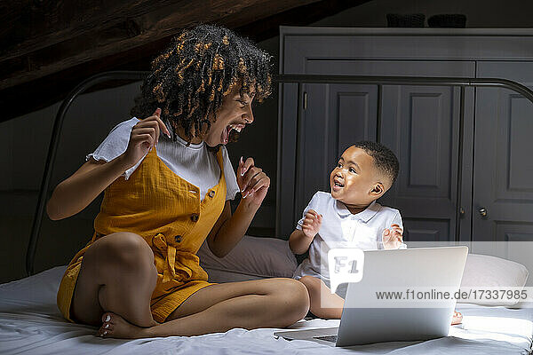 Fröhliche Mutter und Sohn mit Laptop schauen sich gegenseitig an  während sie zu Hause auf dem Bett sitzen