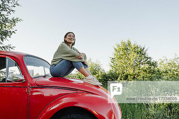 Smiling young woman hugging knees sitting on car hood
