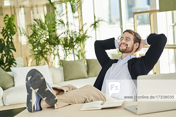 Lächelnder  entspannter männlicher Berufstätiger  der mit hochgelegten Füßen auf dem Tisch im Heimbüro sitzt