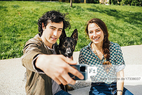 Lächelnder Freund  der ein Selfie mit Hund und Freundin über sein Smartphone auf dem Fußweg macht