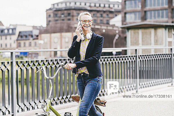 Smiling female commuter wheeling bicycle while talking on mobile phone in city
