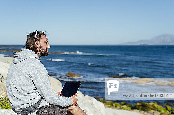Lächelnder männlicher Freiberufler mit Blick auf das Meer  während er mit seinem Laptop am Strand sitzt