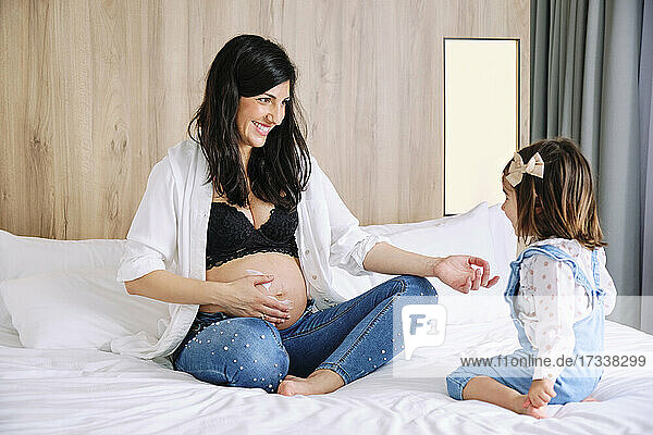 Glückliche schwangere Mutter  die ihre Tochter anschaut  während sie den Bauch mit Feuchtigkeitscreme einreibt