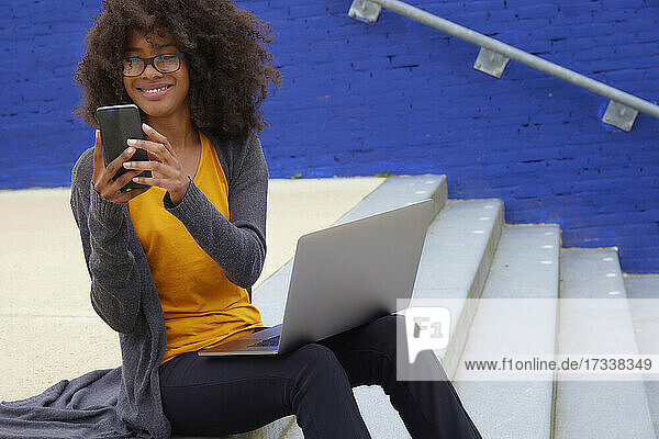 Lächelnde Afro-Frau  die ein Mobiltelefon benutzt  während sie auf einer Treppe sitzt