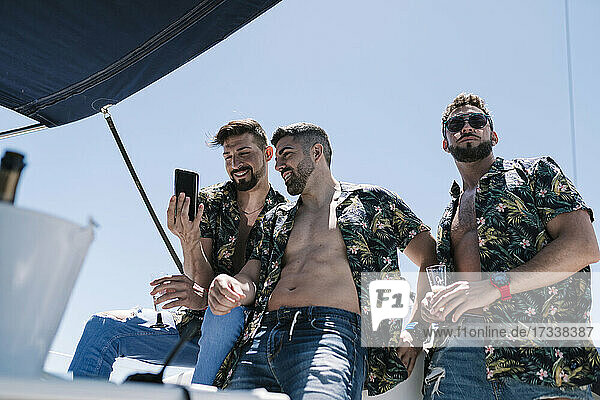 Mann schaut weg  während männliche Freunde auf einer Jacht ihr Handy teilen