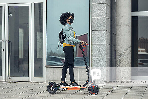 Frau mit Schutzmaske fährt während der COVID-19-Krise mit einem Elektroroller am Gebäude