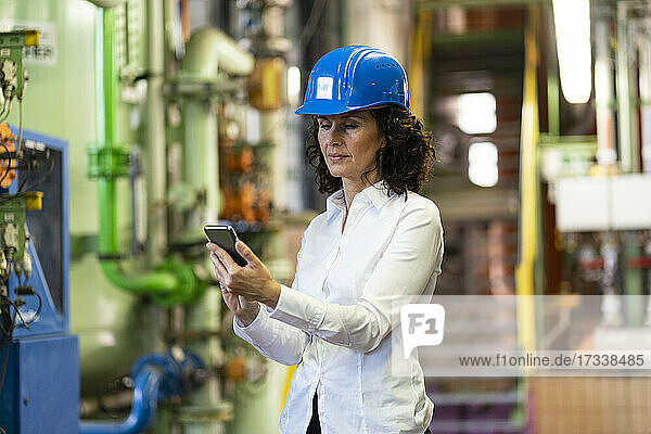 Weiblicher Ingenieur mit Schutzhelm  der ein Mobiltelefon benutzt  während er in der Industrie steht