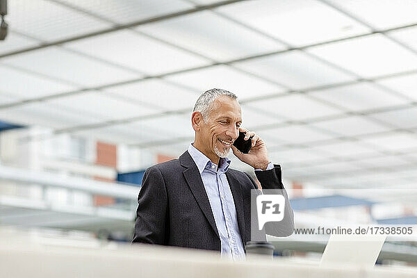 Lächelnder Geschäftsmann  der mit seinem Smartphone telefoniert und auf einen Laptop am Bahnhof schaut