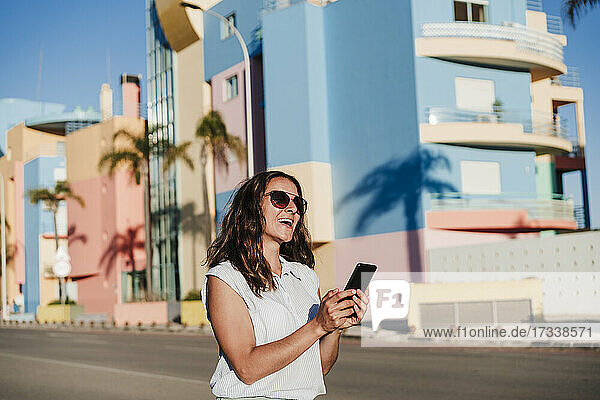 Fröhliche Frau mit Sonnenbrille  die ihr Smartphone hält  während sie auf der Straße in der Stadt steht