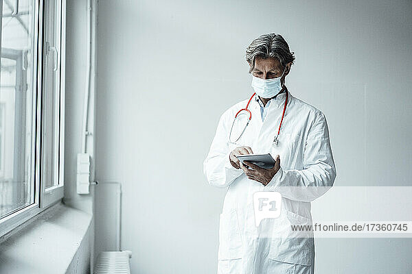 Älterer männlicher Arzt bei der Verwendung eines digitalen Tablets im Krankenhaus während COVID-19