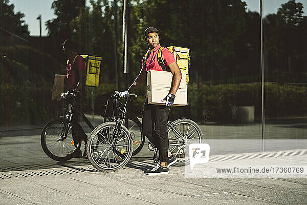 Männlicher Zusteller  der ein Paket trägt  während er mit dem Fahrrad an einer Glaswand steht