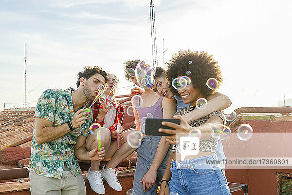 Junge männliche und weibliche Freunde  die ein Selfie machen  während sie auf der Terrasse Seifenblasen blasen