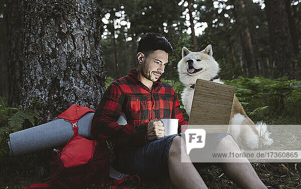 Male hiker working on laptop while sitting by dog in forest during sunset