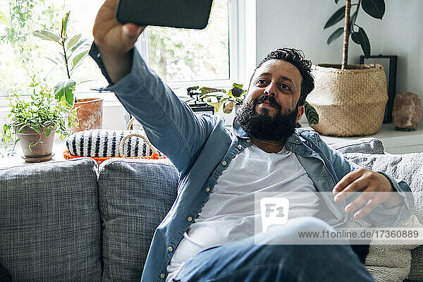 Smiling man taking selfie through smart phone while sitting on sofa at home