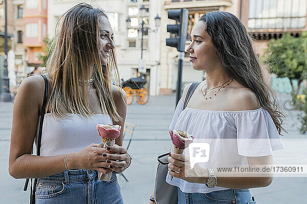 Young women holding ice cream cones while looking at each other in city